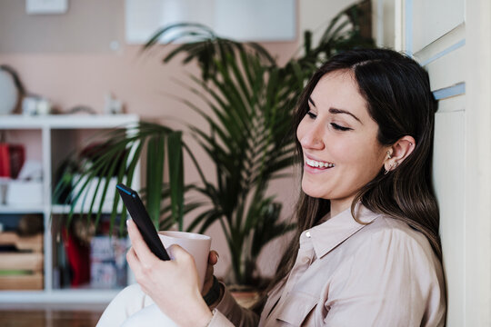 Smiling Mid Adult Woman Using Smart Phone While Holding Coffee Cup At Home