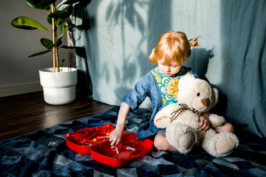 Playful blond girl aspiring doctor while holding teddy bear at home