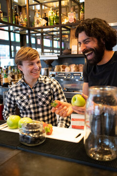 Male bartender laughing while holding fresh fruit by trainee at bar counter