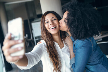 Smiling businesswoman taking selfie while being kissed by female colleague in office