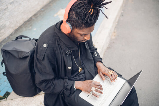 Young freelance worker with headphones using laptop while sitting on footpath