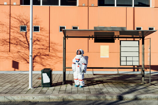 Young female astronaut in space suit waiting at bus stop during sunny day