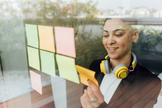 Smiling female entrepreneur sticking adhesive notes on glass in office