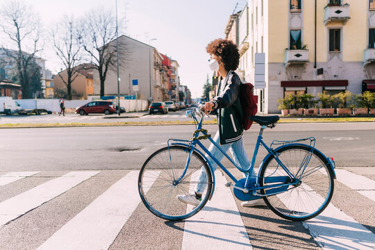 Young woman wearing protective face mask crossing road with bicycle on sunny day