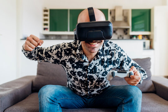 Smiling man wearing virtual reality headset punching while holding game controller on sofa at home