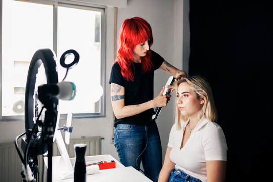 Female hairstylist using straightener on model's hair in front of ring light at studio
