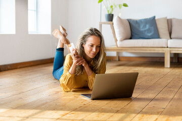 Blond woman with credit card lying down on floor while using laptop at home