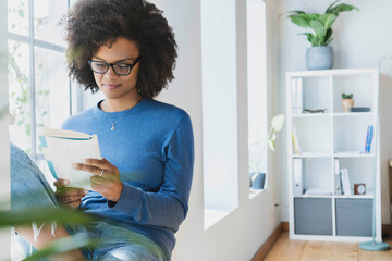 Young woman reading book while sitting by window at home