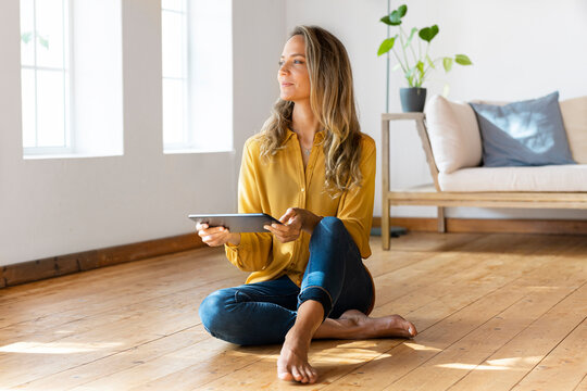 Woman with digital tablet looking away while sitting on floor in living room