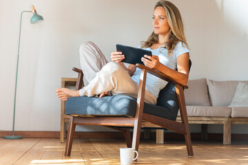 Contemplating woman holding digital tablet while sitting on armchair in living room
