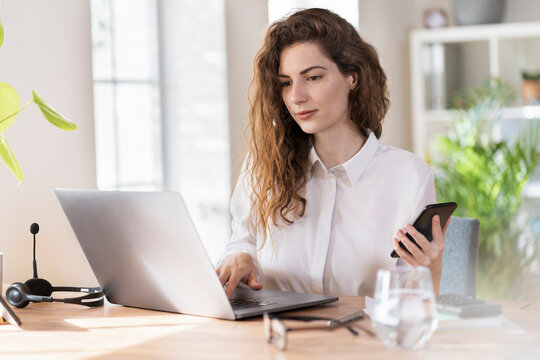 Female Professional With Mobile Phone Working On Laptop At Desk