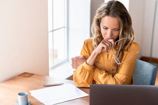 Female professional holding eyeglasses while looking at laptop on desk