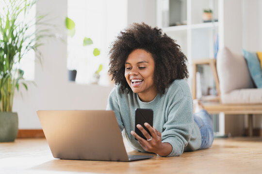 Smiling woman with mobile phone using laptop while lying on floor at home