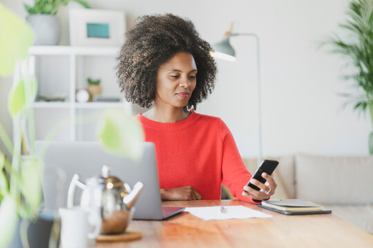 Young woman using mobile phone while sitting by laptop at home