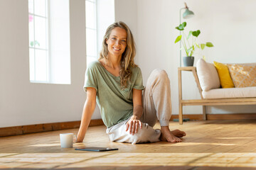 Happy woman sitting on floor by digital tablet and mug at home
