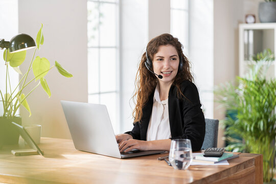 Smiling female customer service representative with headphones looking away while sitting in front of laptop at desk
