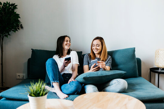 Smiling girlfriends using mobile phones while sitting on sofa at home