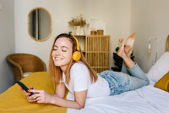 Smiling woman with eyes closed listening music through headphones on bed at home
