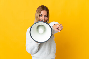 Young blonde woman isolated on yellow background shouting through a megaphone to announce something while pointing to the front