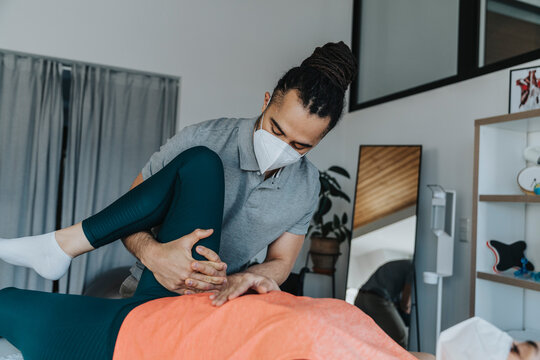 Physiotherapist In Protective Face Mask Doing Therapy On Knee Of Female Patient In Medical Practice During Pandemic