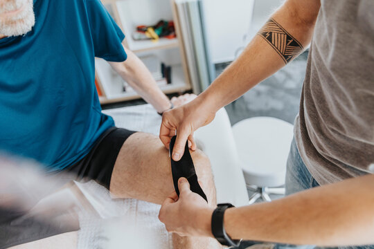 Male physiotherapist sticking elastic therapeutic tape on knee of patient