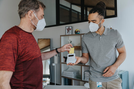 Physiotherapist in protective face mask holding credit card reader while patient paying in medical practice