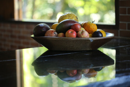 Frutas da esta&ccedil;&atilde;o dentro de uma gamela de madeira, sobre um balc&atilde;o de pedra preta. Ao fundo desfocado,  uma paisagem verde por fora da janela.