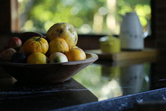 Frutas da esta&ccedil;&atilde;o dentro de uma gamela de madeira, sobre um balc&atilde;o de pedra preta. Ao fundo desfocado, uma garrafa t&eacute;rmica e um a&ccedil;ucareiro e uma paisagem verde por fora da janela.