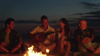 Close-up view of four friends sitting around campfire on beach after sunset and chatting. Man strumming acoustic guitar. Summer evening at seaside. - Powered by Adobe