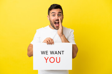 Young handsome caucasian man isolated on yellow background holding We Want You board and shouting