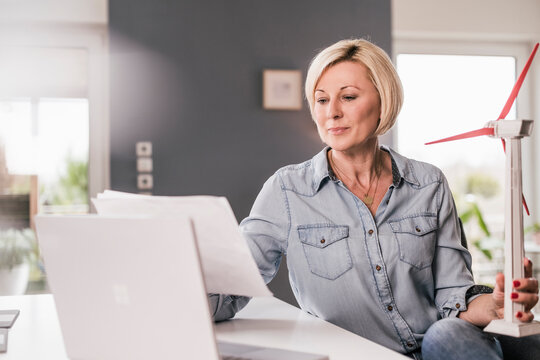 Mature businesswoman reading paper while holding wind turbine model at home office