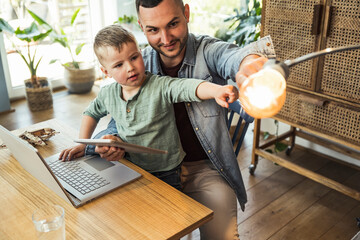 Man and son pointing at light bulb while sitting with tablet and laptop in smart home