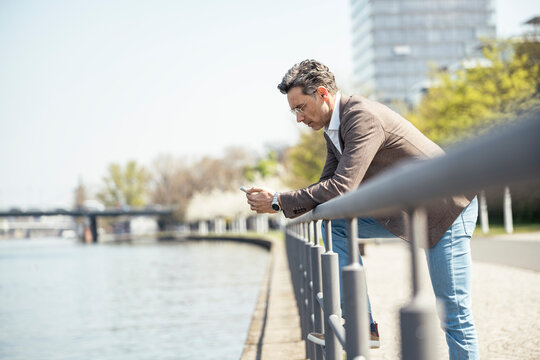 Mature businessman using mobile phone while leaning on railing by river