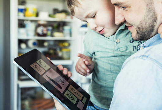 Smiling mid adult man carrying son pointing at digital tablet in kitchen