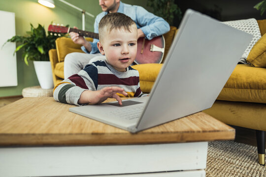 Cute boy using laptop while father playing guitar in living room