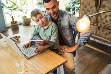 Father and son looking at light bulb while sitting with tablet and laptop in smart home