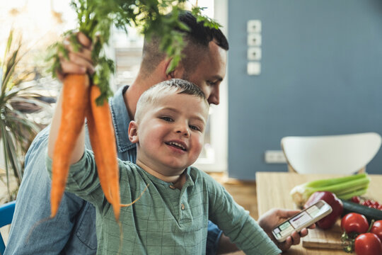 Boy holding carrots while father using smart phone at home