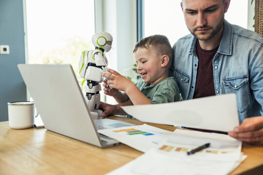 Smiling boy playing with robot while businessman reading documents at home