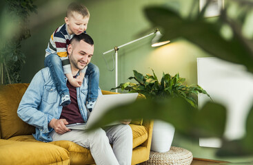 Smiling man using laptop while son sitting on shoulder in living room at home