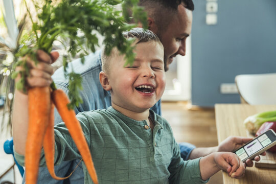 Smiling boy with eyes closed holding carrots while father using mobile phone at home