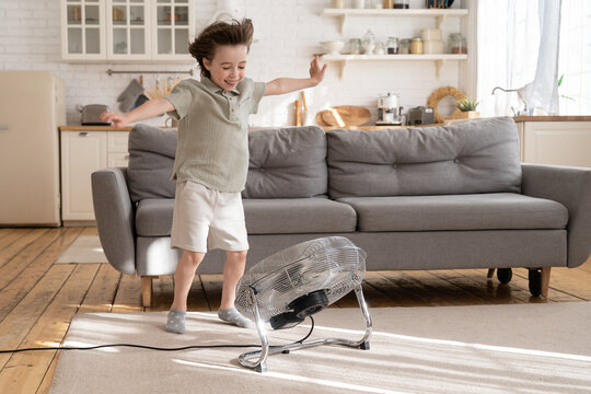 Little Boy Play With Strong Wind Blow From Ventilator Or Industrial Fan At Home In Cozy Living Room. Small Kid Alone With Retro Air Conditioner Enjoy Refreshing Fresh Breeze. Child Has Fun With Cooler