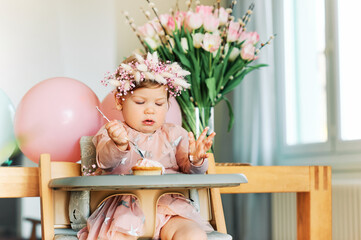 Adorable 1 year old baby girl eating cupcake, happy child sitting in a chair, tasting sweet dessert, first birthday