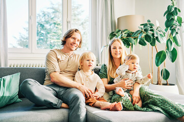 Happy young family of four relaxing on a couch, couple playing with baby girl and toddler boy,...