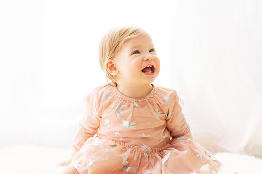 Light Soft Portrait Of Cute Toddler 1 Year Old Baby Girl, Wearing Pink Dress, Sitting Next To Window