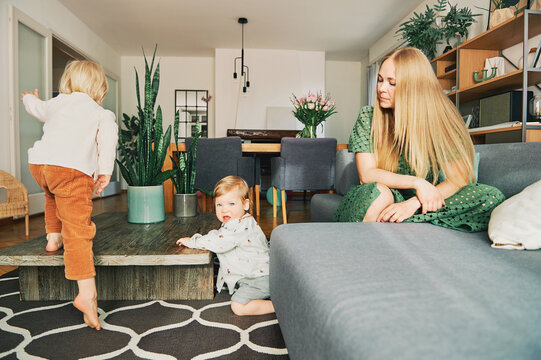 Young Mother Resting On Couch While Little Kids Playing And Jumping In Living Room