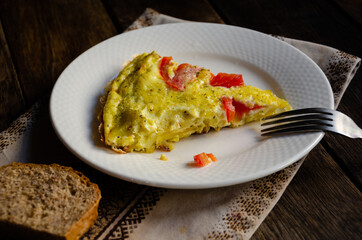 A serving of rustic omelet in a white plate, bread, kitchen napkin on a wooden table.