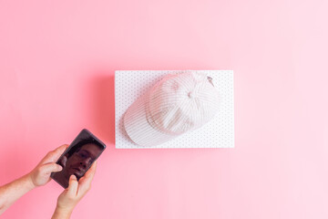 Obraz na płótnie Canvas top view of box with cap inside on pink background with man reflected on smartphone screen. concept buying clothes online. electronic commerce. flat lay flat design