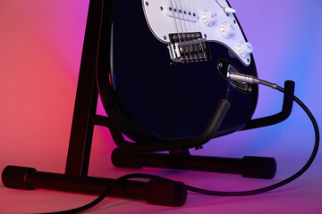 An electric guitar on a stand connected with a cord to an amplifier is illuminated with blue and...