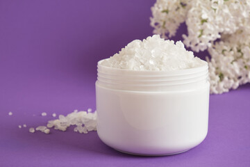white sea salt in plastic jar and white lilac flowers on lilac background