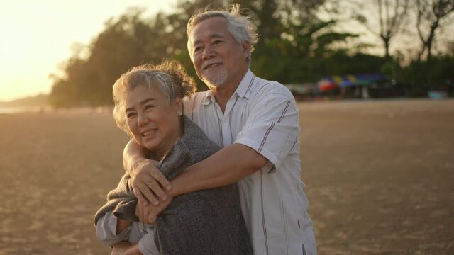 Happy Asian family senior husband cover his wife with a blanket and hugging together on the beach at summer sunset. Healthy elderly retirement couple relax and enjoy romantic outdoor vacation together